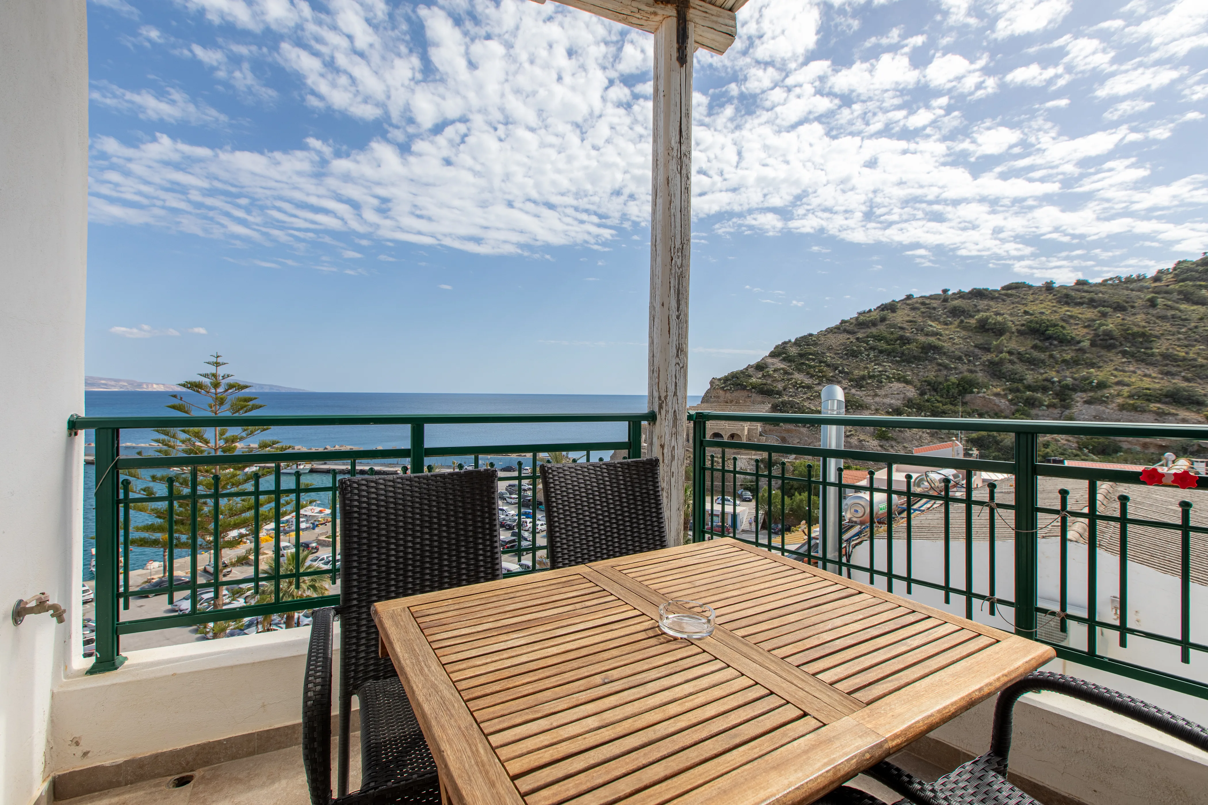 outside view of the apartment's balcony with a wooden table with armchairs and the view of the sea and a cliff at the background during the day
