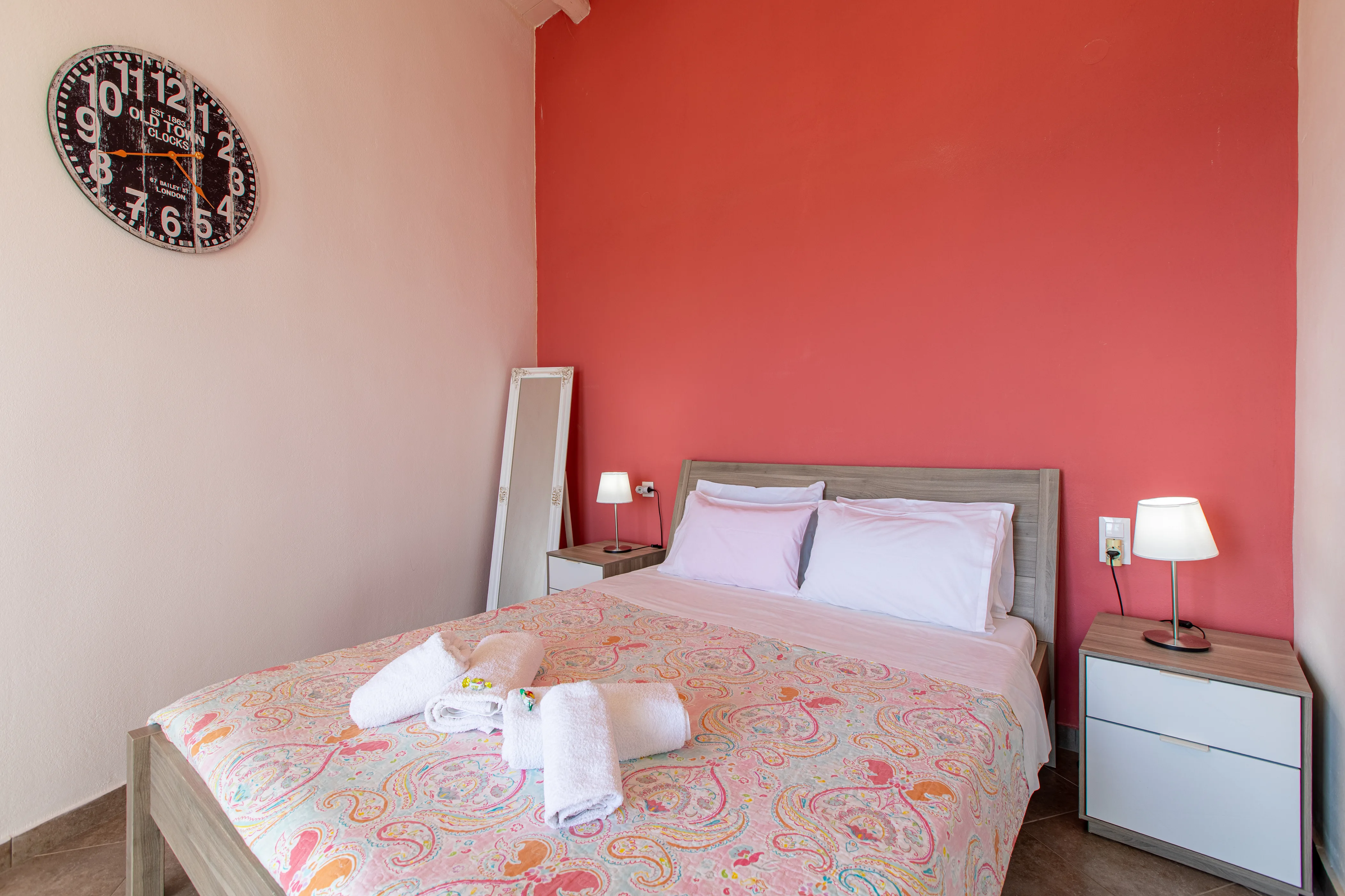 inside view of the apartment room featuring a large double bed from the front side with a red painted wall behind it and a nightstand on the right side of the bed
