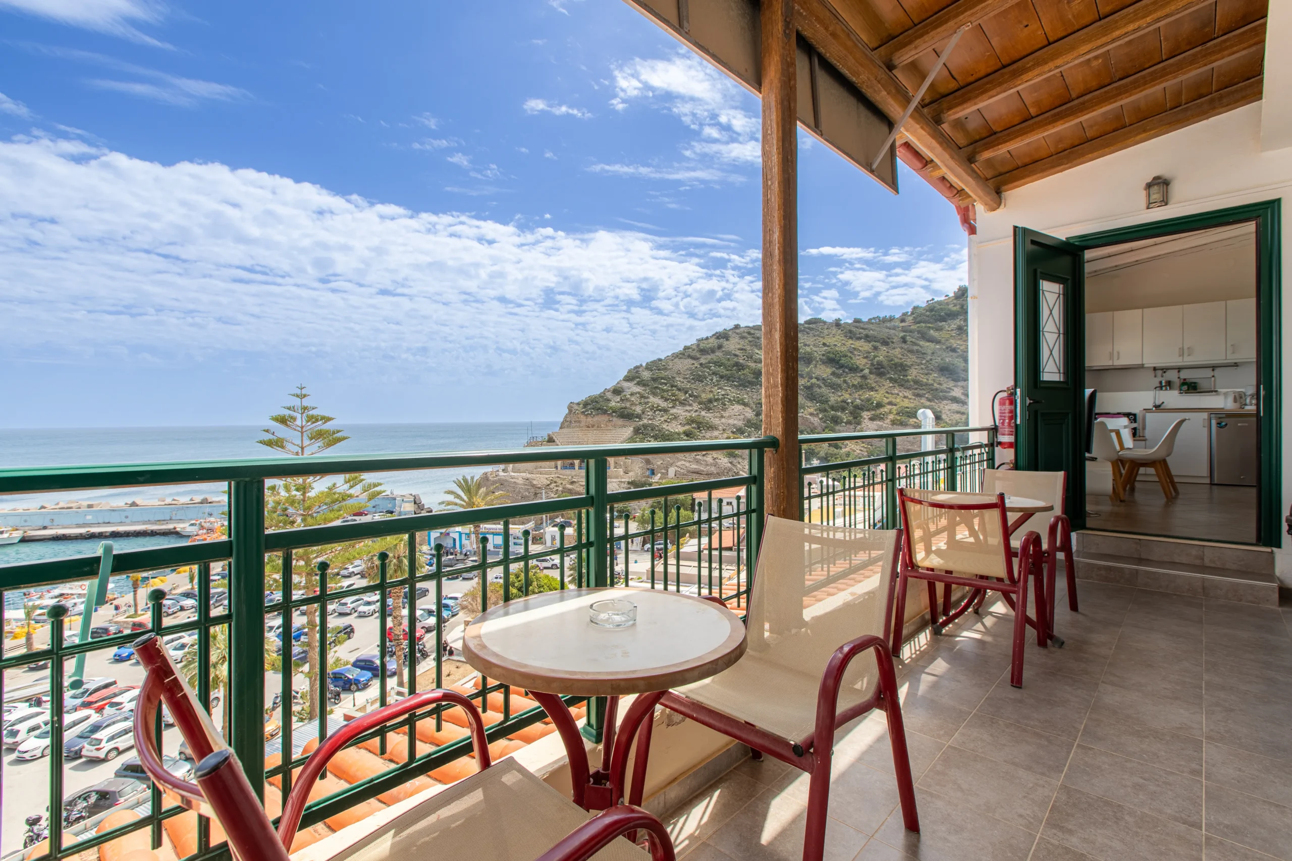 realistic view from the hotel balcony featuring some tables with armchairs and the sea at the background and a opened room door on the end of the balcony