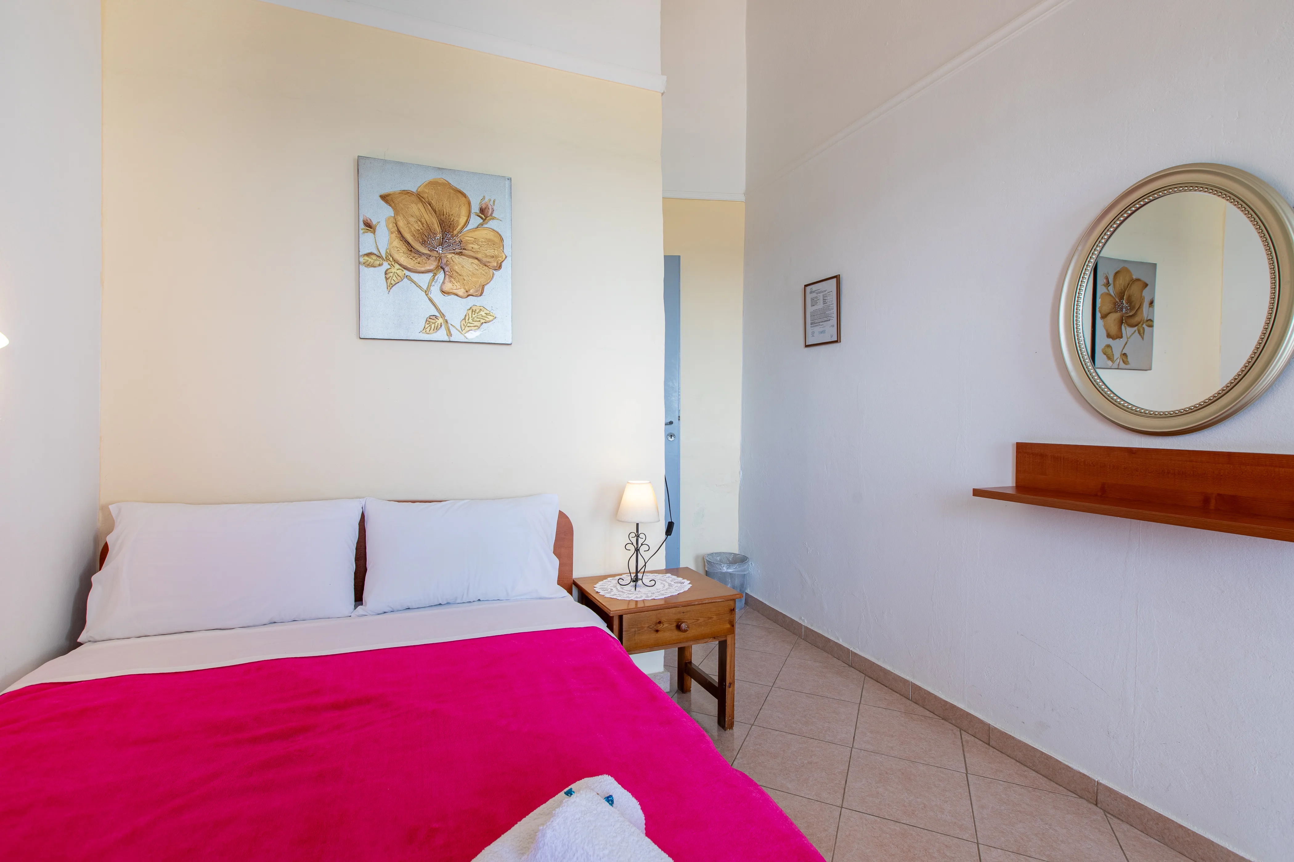 inside view of a room featuring a double bed with red bedsheets and a wooden nightstand next to it and a mirror on the right side wall